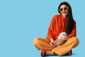Young happy woman in sunglasses sitting with her cute Bolognese dog on blue background
