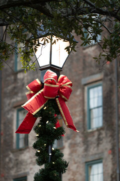 A street lamp decorated for Christmas with a red and gold bow, green garland, and yellow Christmas lights, with brick apartment building in background