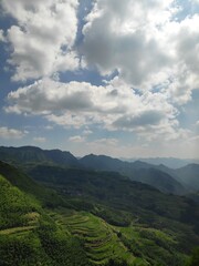 Naklejka premium This breathtaking image captures a panoramic view of expansive mountain ranges with lush, terraced fields below under a beautiful sky dotted with fluffy white clouds.