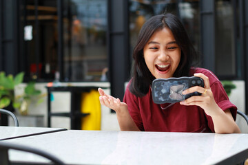 attractive asian woman excited playing game online using smartphone with raised hand sitting in outdoor cafe