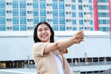 Fototapeta premium beautiful asian young woman holding sparklers firework and laughing in new year eve celebration standing in roof top outdoor with urban building background