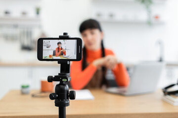 Young woman recording video blog at home on smartphone. She sits at desk with laptop and coffee cup, focusing on creating content.