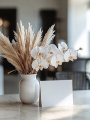 A white vase with pampas grass and white orchids, and a blank white card on a marble table in a modern setting.