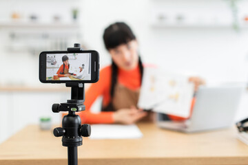 Caucasian woman influencer in her 20s creates engaging video content in bright home environment. Seated at table, she presents material in front of smartphone camera on tripod.