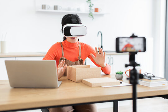 Caucasian female blogger unboxing new virtual reality headset for online audience. Young woman sharing engaging tech content from home office. Creative setup with laptop, smartphone, and modern desk.