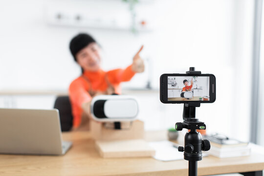 Caucasian female blogger unboxing virtual reality headset on camera. She is seated at desk with laptop, creating engaging online content. Setting includes phone recording video of unboxing session.