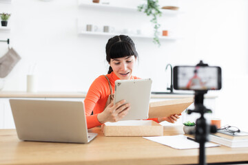 Caucasian young woman creating online content while unboxing new tablet. Modern bright kitchen setting enhances vibrant atmosphere. Enthusiastic expression conveys excitement about new technology.