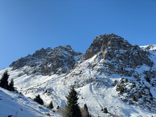 A stunning view of the rugged mountain peaks in Almaty, Kazakhstan. The golden sunlight casts a warm glow on the rocky formations, contrasting beautifully with the snow-covered slopes below.