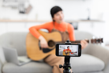 Caucasian woman recording online acoustic guitar course at home. Seated on sofa, she plays guitar in front of camera while laptop is nearby. Focus is on creative content creation, teaching, and music.