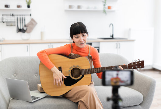 Caucasian woman recording online guitar lesson at home. She sits on couch with acoustic guitar, laptop, and camera. Casual and creative ambiance perfect for music and education.