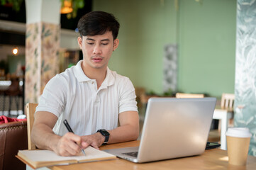 Fototapeta premium A serious, focused Asian man is working remotely from a coffee shop, taking notes in his notebook.