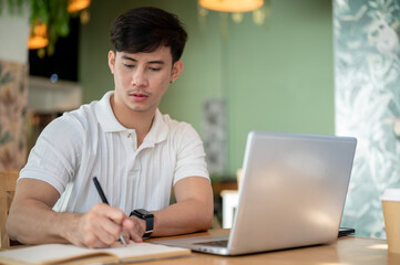 A serious, focused Asian man is working remotely from a coffee shop, taking notes in his notebook.