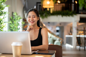 An Asian woman sitting at an indoor table in a coffee shop with her laptop, smiling at the camera.