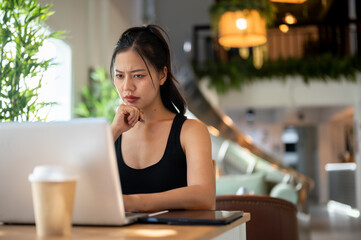 A stressed Asian woman reading something intently on her laptop with a serious expression.