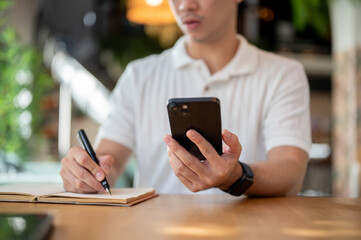 A cropped shot of an Asian man holding his smartphone while taking notes in his notebook.