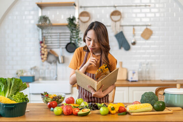 Asian young woman holds a book with online audience or simply enjoying the cooking process, health, fruit, freshness, and organic living, digital recipe platform, eating, healthy at kitchen