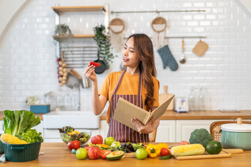 Asian young woman holds a book with online audience or simply enjoying the cooking process, health, fruit, freshness, and organic living, digital recipe platform, eating, healthy at kitchen