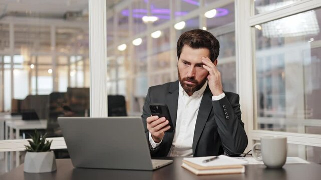 Stressed businessman having a headache while reading bad news on his phone. Male manager frustrated, angry, upset, worried about message big problem at work. Shocked man in panic at office workplace.