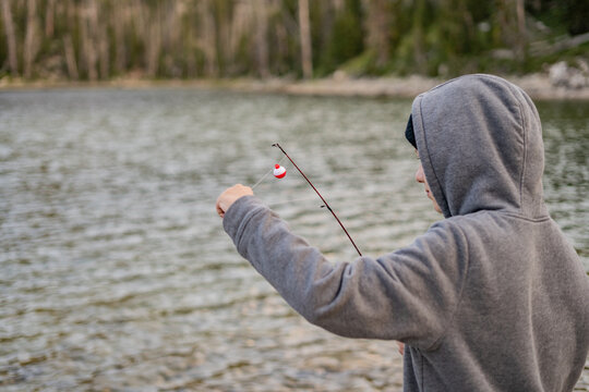 Person in hoodie fishing by lakeside holding fishing pole