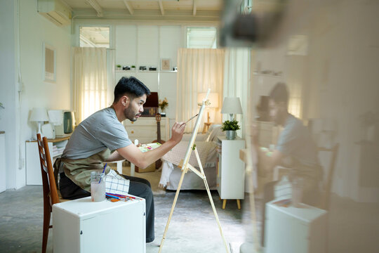 Young Asian male artist in apron carefully works with watercolor painting on paper with intense focus in a peaceful home studio environment, hobby activity concept.