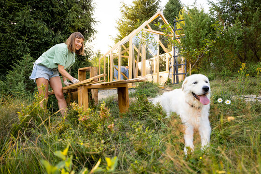 Female carpenter standing by a workbench and her large white dog in front of a partially built wooden greenhouse in her backyard. Engaged in a DIY project, surrounded by nature
