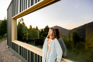 A woman leans against a modern cabin, reflecting on the tranquility of nature surrounding her. The...