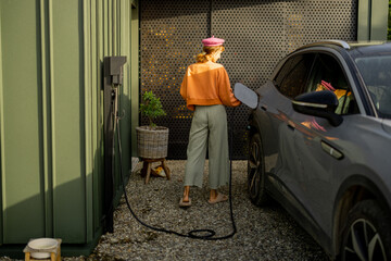 A woman in a stylish outfit charges her electric car at a modern home nestled in nature, highlighting a blend of sustainable living and contemporary lifestyle