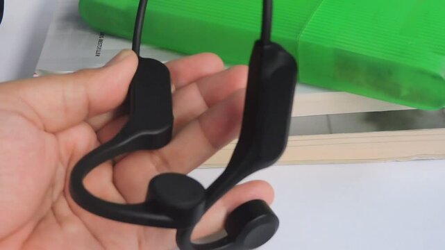 a hand holds a pair of sleek black wireless bone conduction headphones next to a smartphone, highlighting its compact and ergonomic design. 