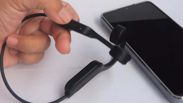 a hand holds a pair of sleek black wireless bone conduction headphones next to a smartphone, highlighting its compact and ergonomic design.