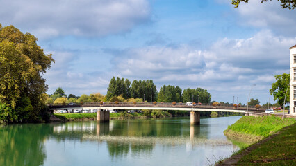 Peyrehorade, France, the bridge spans a river with a green reflection on the water. The bridge is surrounded by trees and there are cars