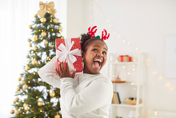 Portrait of excited young happy african american woman holding red present gift box standing in living room at home. Joyful girl celebrating New Year near fir tree. Christmas celebration concept.