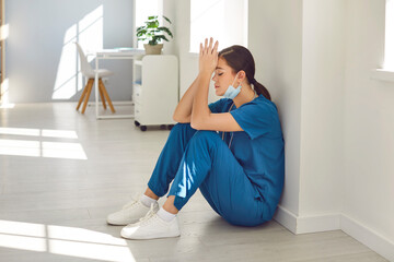 Exhausted stressed female nurse or doctor wearing blue face mask and uniform sitting with closed eyes on hospital floor in hallway. Young woman physician tired from hard work in medical clinic.