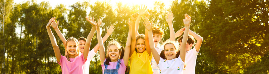 Group of smiling kids friends raising hands up on green grass in the park standing in a line. Happy children having fun together outdoors on a sunny summer day in casual clothes in the camp. Banner.