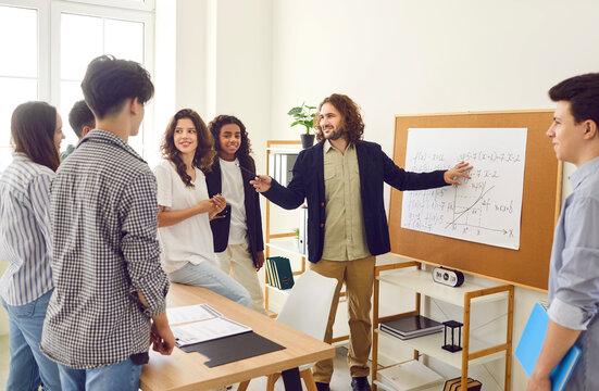 Teacher and teenage students having math class at school. Friendly male teacher standing by board in classroom and explaining algebraic functions to group of adolescent schoolgirls and schoolboys
