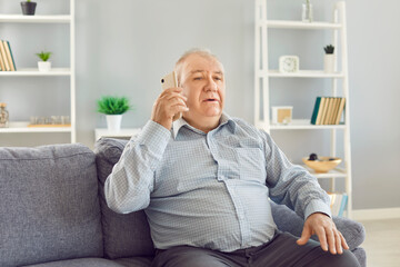 Fototapeta premium Serious elderly man sitting on a sofa in the living room, engaged in a phone call an talking. His deep conversation, highlighting the importance of communication in home environment.