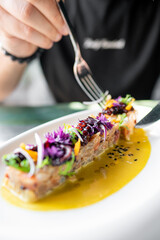 A hand holding a fork, poised to enjoy a gourmet dish garnished with edible flowers and herbs, presented on an elongated plate with a vibrant sauce. The setting is elegant, highlighting fine dining