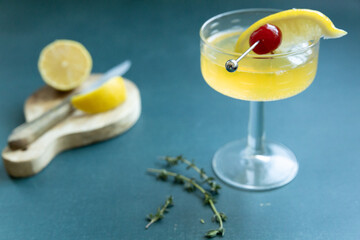 Yellow cocktail in glass with lemon and cherry garnish on a table
