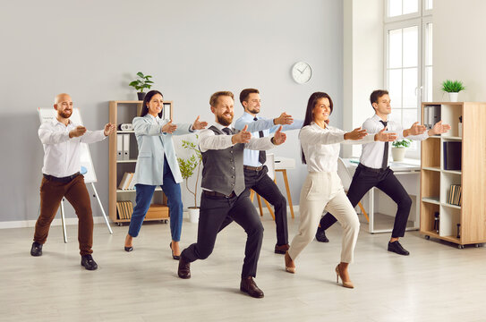 Full length portrait of young funny business people doing corporate exercises standing at workplace in office. Team of smiling company employees exercising for health during a break from a work.