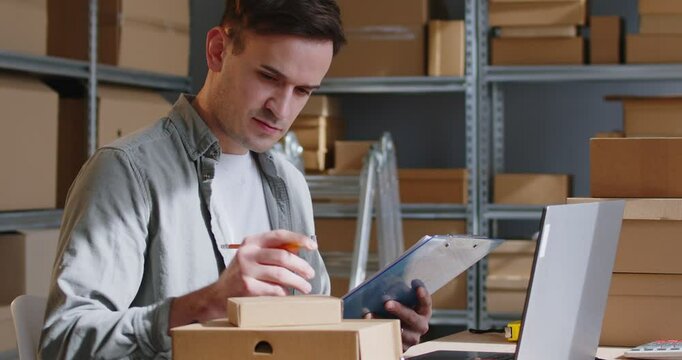 Man working inventory online store warehouse cross referencing boxes with checklist. Attention to detail of goods inspection aspect of online store warehouse operations at warehouse.Post office.
