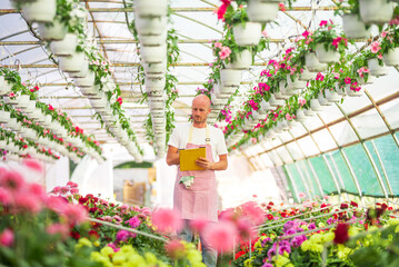 Gardener checking flowers standing in greenhouse holding clipboard
