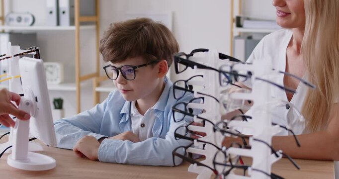 Nice little happy child boy and his mother choosing glasses at optician store or in clinic with doctor ophthalmologist consultation. Vision correction and ophthalmology concept. 4k video.