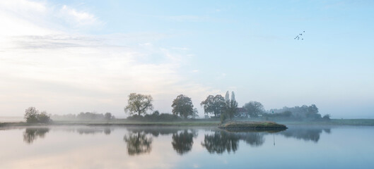 Naklejka premium farmhouse reflected in water of river lek near wijk bij duurstede in holland on foggy morning