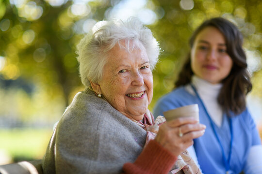 Elderly woman enjoying cup of coffee with her nurse in the park during warm autumn day. Young caregiver spending time with senior woman.