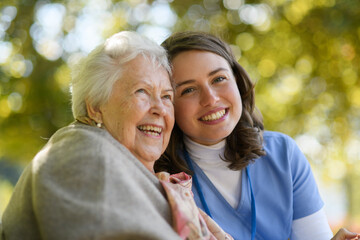 Portrait of nurse and elderly woman on walk in the park during warm autumn day. Young caregiver...