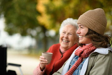 Portrait of a granddaughter and grandmother sitting on bench in autumn park, drinking coffee and...