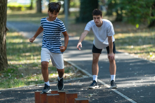 Young boy navigates wooden stumps outdoors under the supervision of a trainer, engaging in balance focused physical activity. The scene highlights childrens outdoor sports activities and exercise.