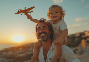 Dad and son with toy plane at sunset. A dad carries his smiling son on his shoulders, who joyfully holds a toy airplane against a sunset.
