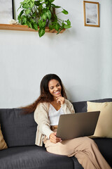 A cheerful woman sits on a soft couch, smiling while using her laptop in a warm and inviting space.