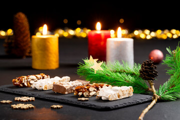 Assortment of gourmet nougat and nut bars on black slate plate under holiday ambiance with candles and evergreen sprigs, pinecone golden star with warm blurred lights perfect for celebrating Christmas