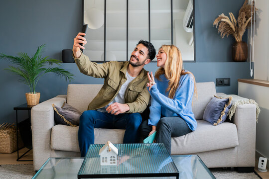 Happy young couple taking a selfie together in their new apartment, sitting on a cozy couch in a stylish living room, celebrating a fresh start and capturing memories in their modern home - Powered by Adobe